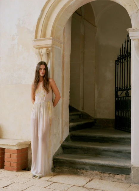 Woman in a long dress poses against a stone archway with an iron gate in the background, soft lighting, neutral colors, elegant pose.