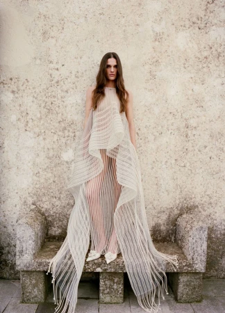 Model wearing a sheer textured white gown standing in front of a weathered stone bench and rustic wall