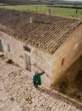 Model wearing a green coat standing on a stone courtyard beside a traditional stone building with a tiled roof.