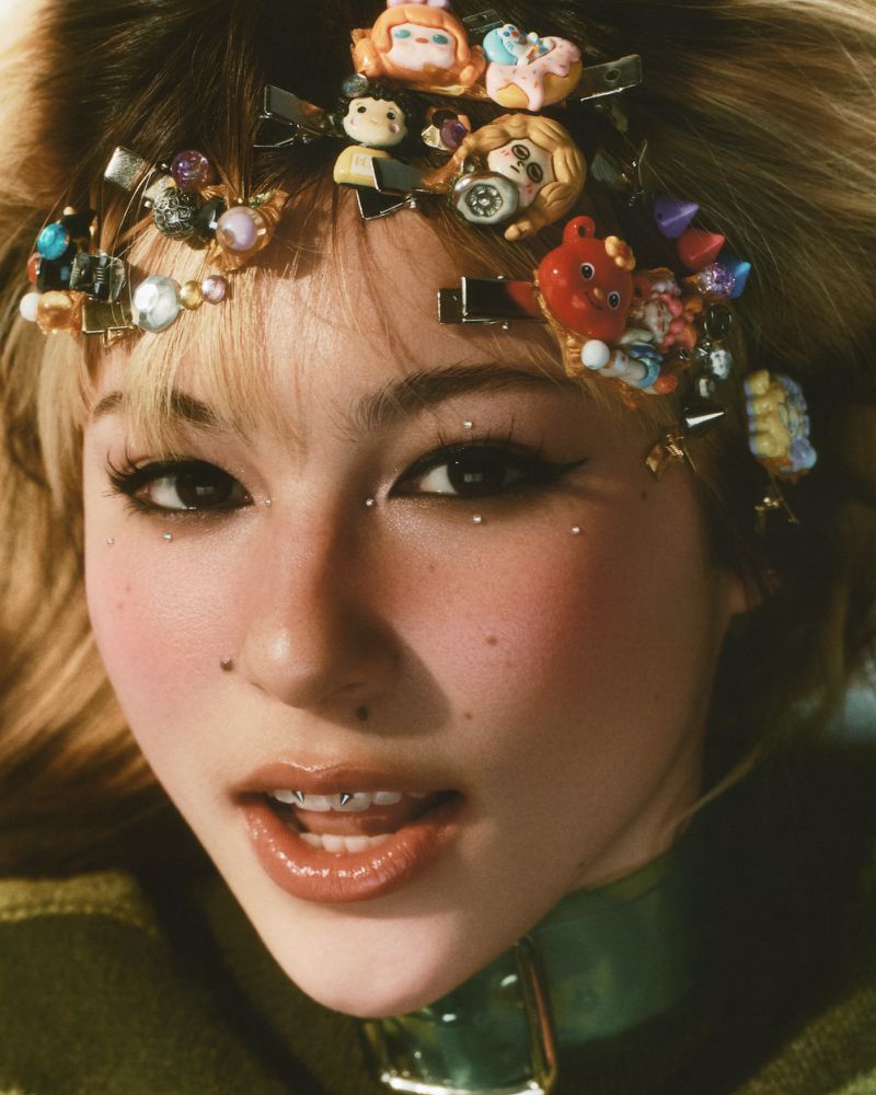 Close-up portrait of a young adult female with face jewelry, colorful hair clips, and olive green clothing, against a neutral backdrop.