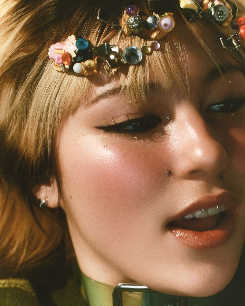 Close-up portrait of a young adult female with blonde-streaked hair, wearing decorative hair accessories and a green choker necklace.