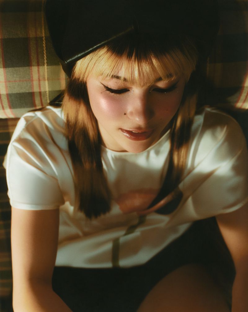 Overhead portrait of a young woman with blond hair and bangs wearing a black hat and white top indoors with warm lighting.