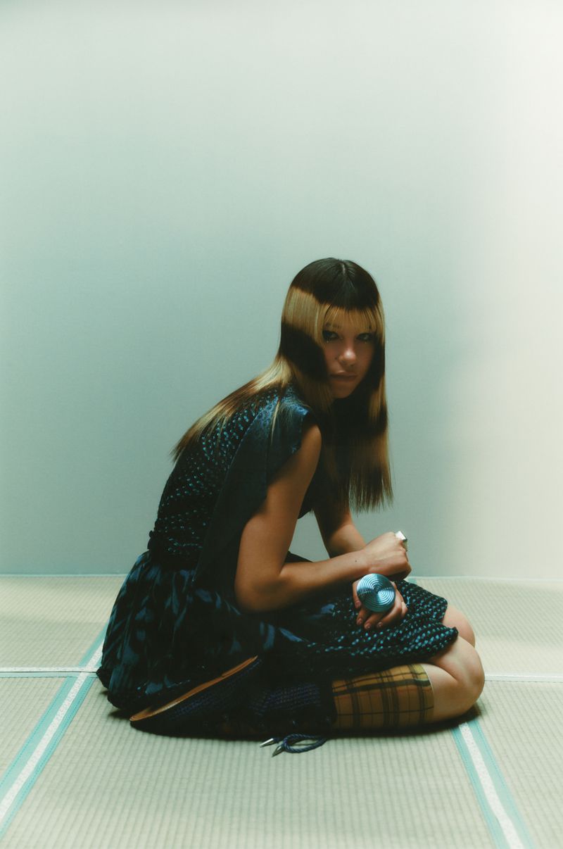 A woman with two-tone hair kneels on a striped mat indoors, holding a round object, wearing a dark dress and a plaid skirt, looking at the camera.