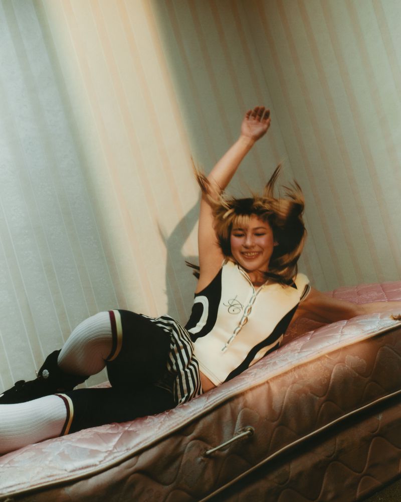 A young woman in sporty outfit lying on a mattress, smiling with her arm raised. Light shines down in an indoor setting.
