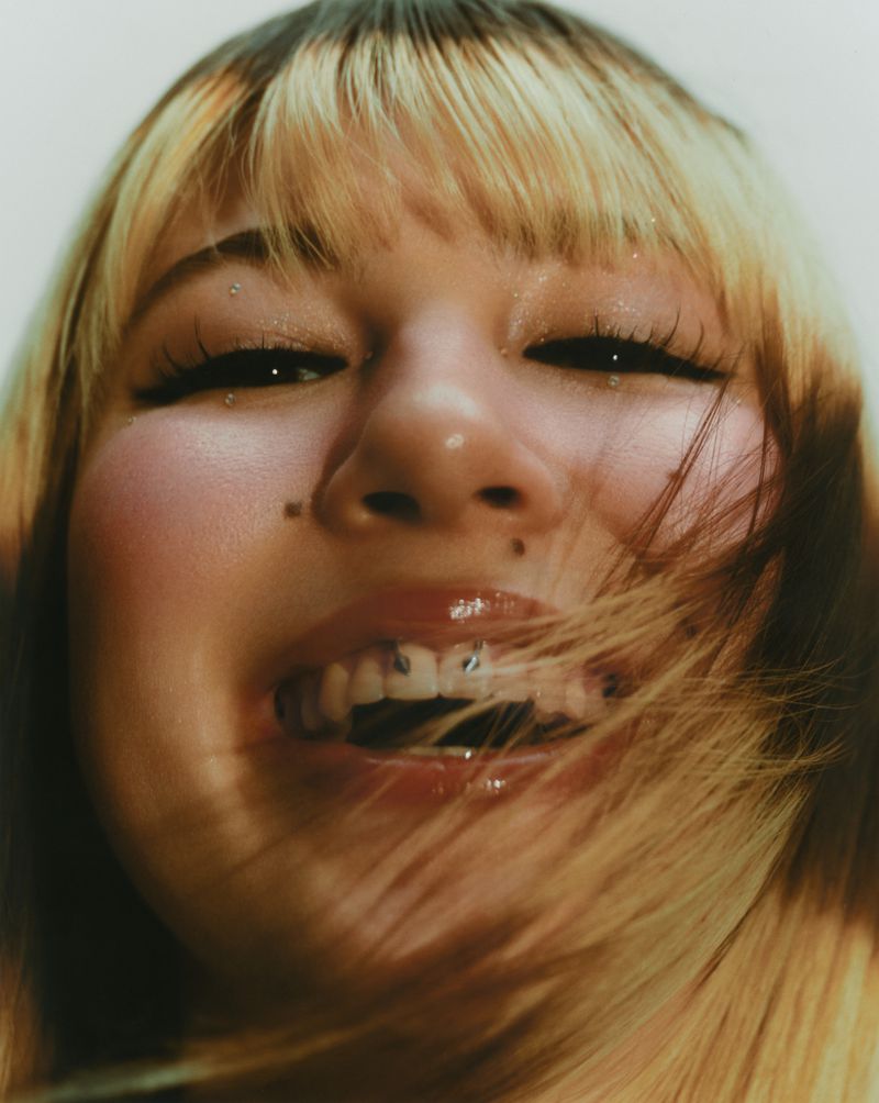 Close-up of a young woman with blonde hair and lip and teeth piercings, smiling and looking directly at the camera in a bright, soft setting.
