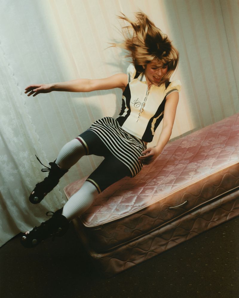 Young woman in motion wearing roller skates, striped skirt, and top with a bicycle design, indoors with pink mattress and white curtain.