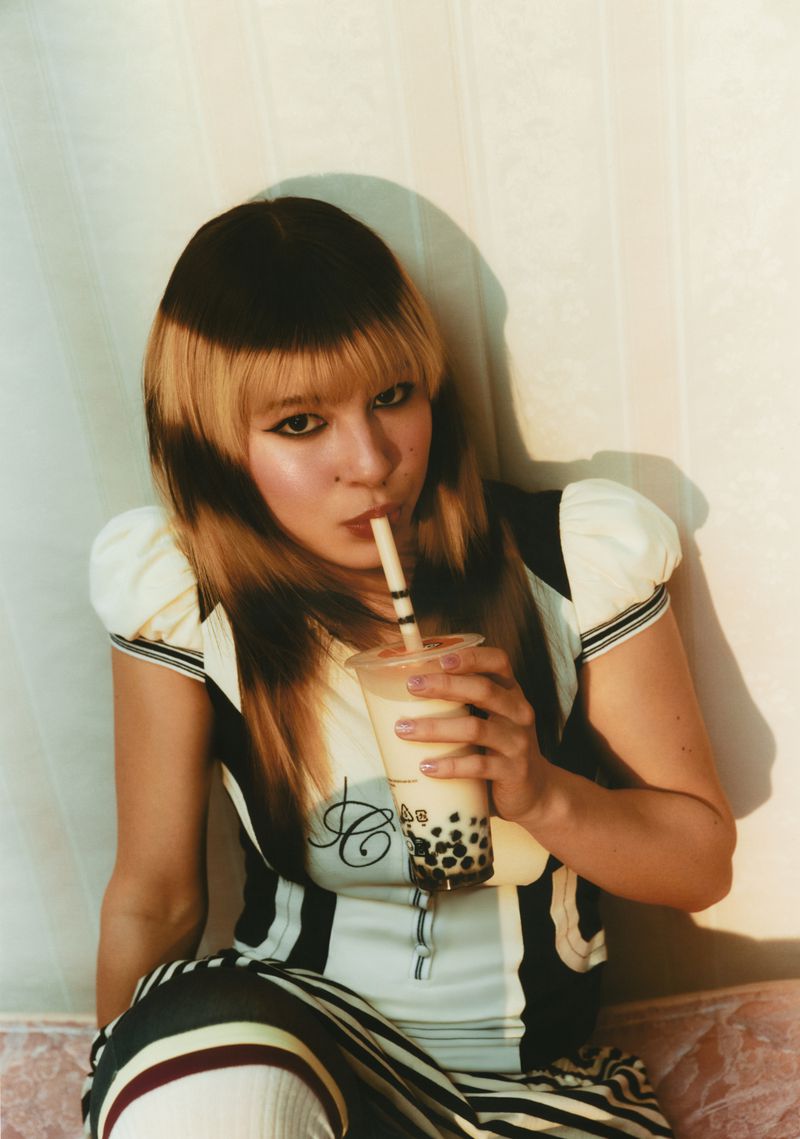 Young woman sips bubble tea in an indoor setting, featuring a black and blonde hairstyle, casual striped attire, and natural lighting.