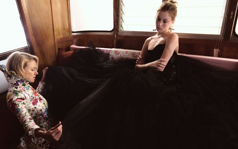 Two blonde women sitting in a wooden interior; one wears a long-sleeved floral jacket, the other a strapless black sequined ballgown.