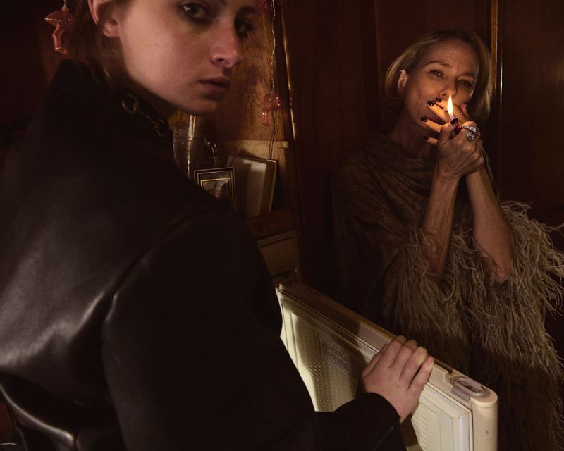 Two women indoors in dim light, one with a lit cigarette, dressed in dark clothing, with a vintage radiator in the foreground.