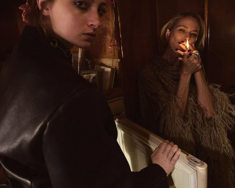 Two women indoors in dim light, one with a lit cigarette, dressed in dark clothing, with a vintage radiator in the foreground.