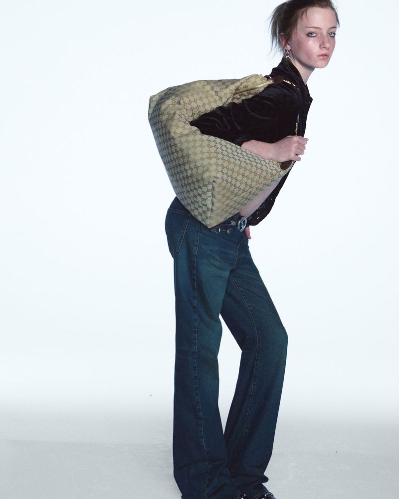 A slender, young woman is posing indoors in a dark jacket, wide-legged jeans and a patterned shoulder bag against a white backdrop.