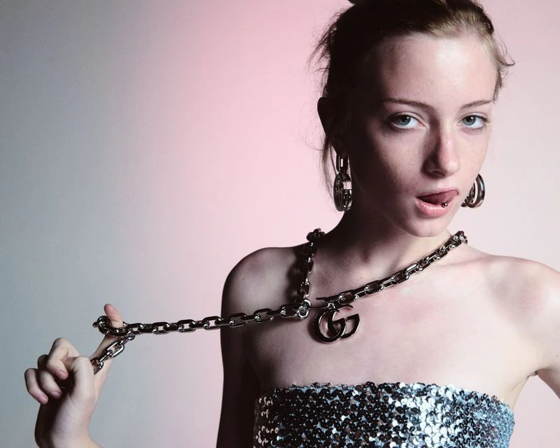 Young woman in silver sequin top poses with heavy metal Gucci chain necklace and hoop earrings against a soft pink background.
