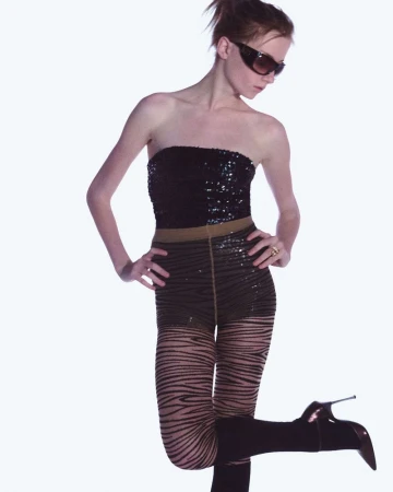 Fashionable woman in studio posing in a black sequin top, patterned tights, and high-heeled boots against a white backdrop. 