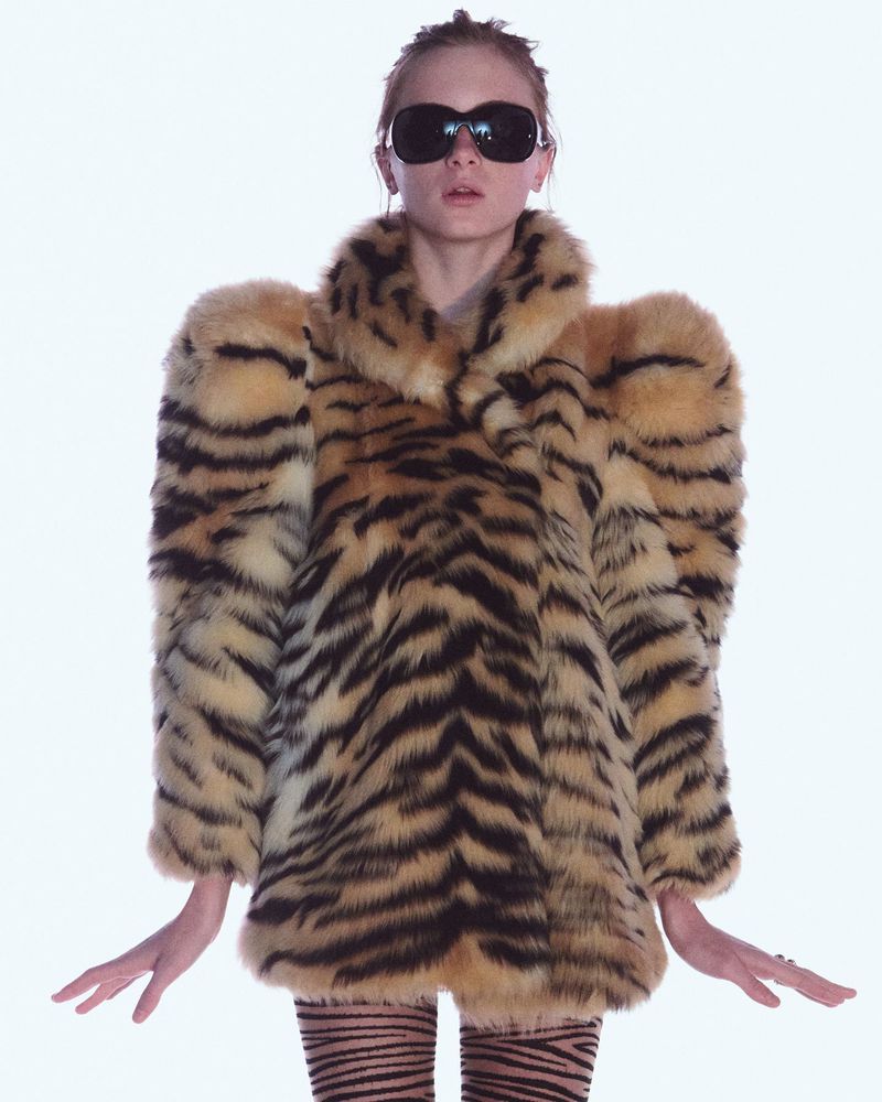 Fashionable adult female poses in a striking tiger-striped faux fur coat and matching leggings against a bright white studio backdrop.