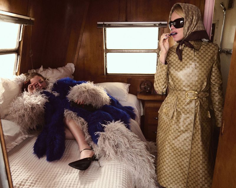 Two women in a vintage camper: one lounges on a bed with a fur blanket, while the other wears a patterned trench coat and headscarf.