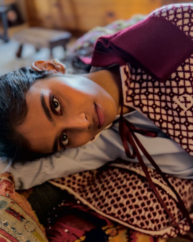 Portrait of a young woman with dark hair laying on a bed with a patterned blanket, wearing a white top and a maroon cardigan.
