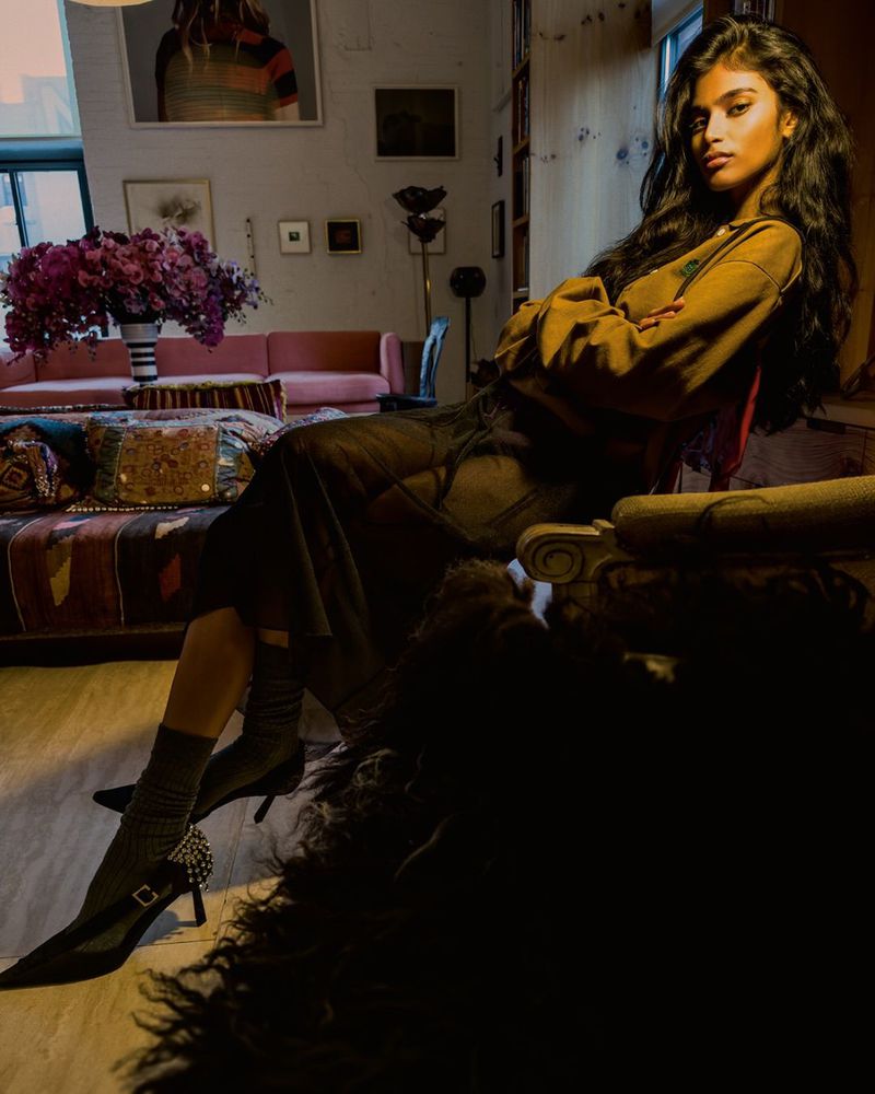 A stylish young woman poses indoors with arms crossed, wearing a brown sweater, a sheer black dress, and heeled sock boots.