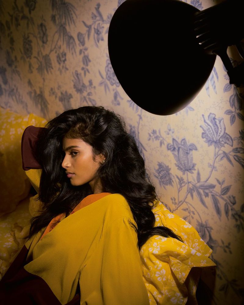 Portrait of a woman in a yellow and orange top reclining against a floral wallpaper background, illuminated by a black lamp.