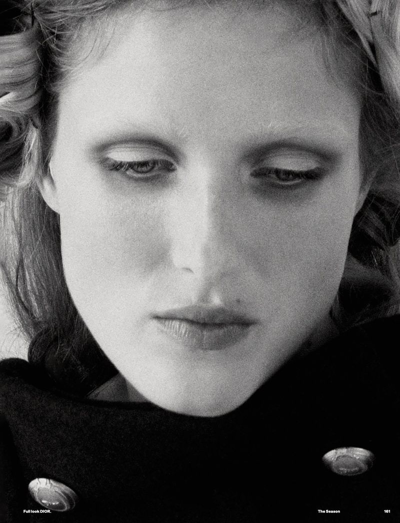 Black and white portrait of a woman with hair rollers, wearing a dark coat with buttons, and looking down in a studio setting.