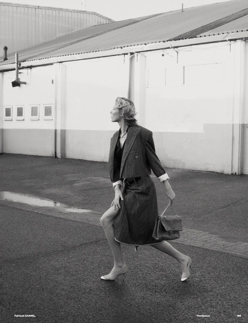 Fashionable woman in coat and skirt walking in urban setting, posing outdoors with purse on wet pavement by building.