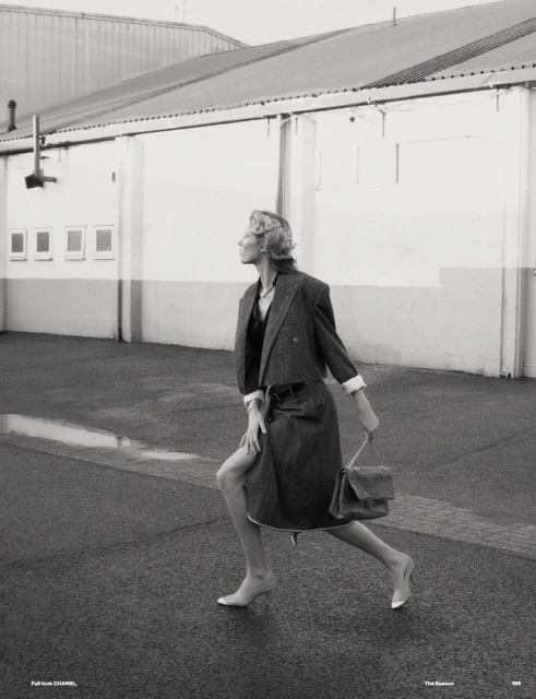 Fashionable woman in coat and skirt walking in urban setting, posing outdoors with purse on wet pavement by building.