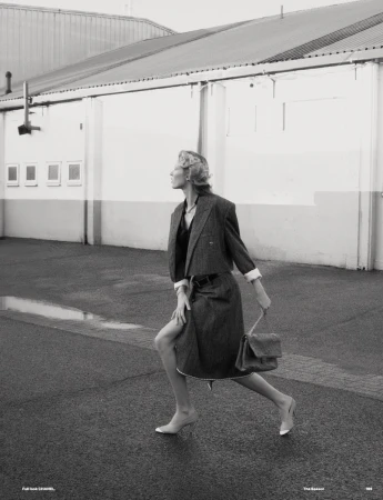 Fashionable woman in coat and skirt walking in urban setting, posing outdoors with purse on wet pavement by building.