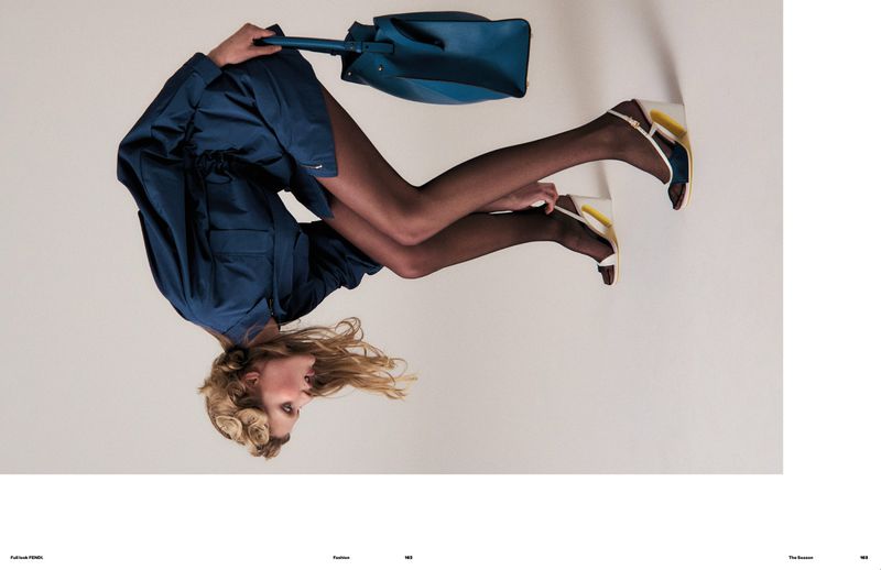 Fashion editorial image of a blonde adult model in a blue dress, black stockings, and white wedge heels, holding a blue purse.