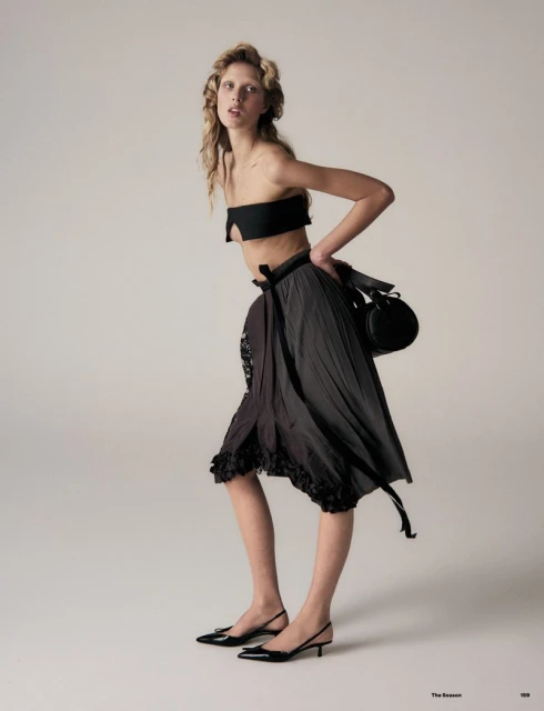 A blonde woman poses in a black bandeau top, grey skirt, and black kitten heels, holding a round handbag against a gradient background.
