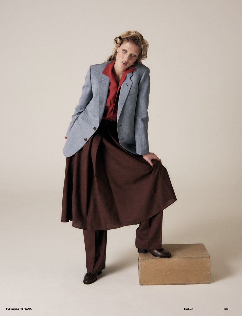 A woman in a red shirt, blue blazer, and brown skirt poses in a fashion shot with a neutral backdrop and a cardboard box prop.