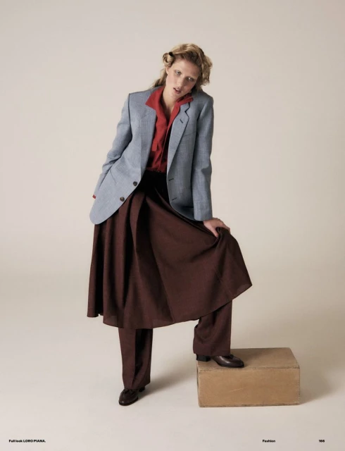A woman in a red shirt, blue blazer, and brown skirt poses in a fashion shot with a neutral backdrop and a cardboard box prop.