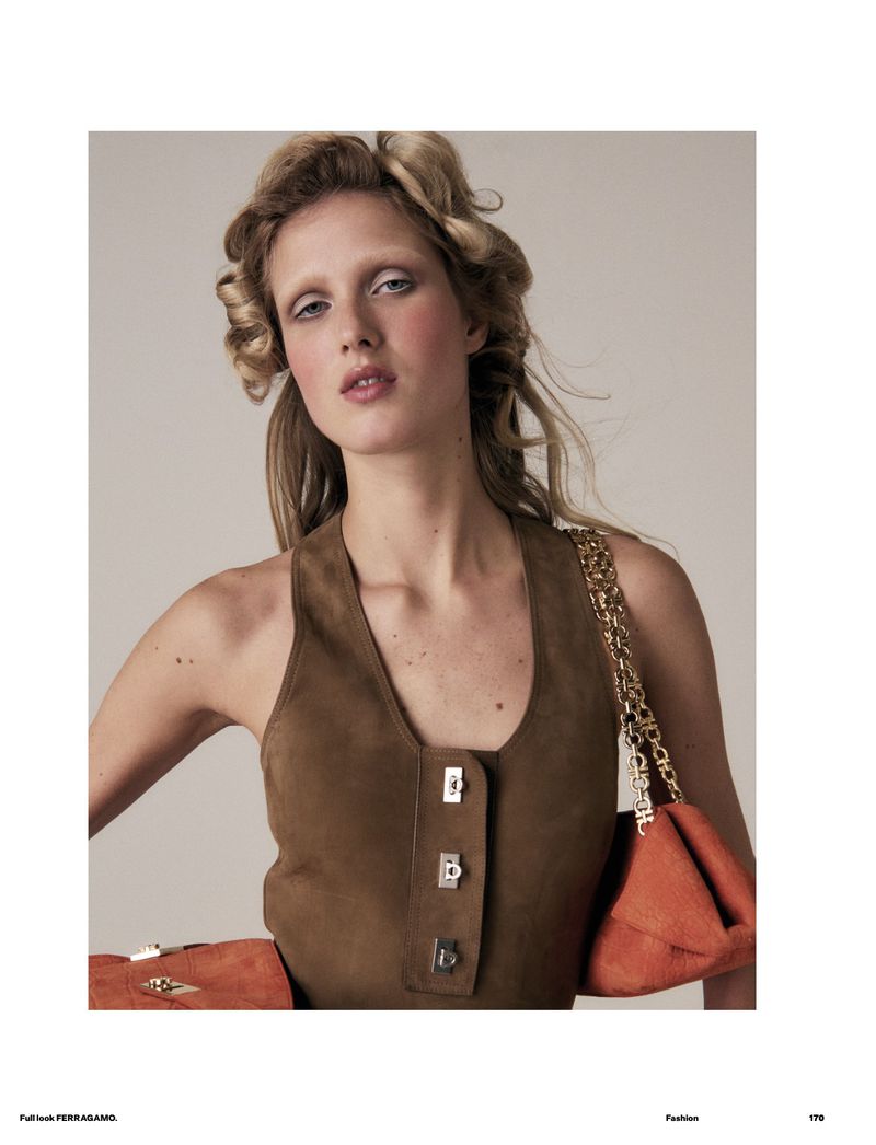 Fashion editorial shot of a fair-skinned woman with blonde hair wearing a brown vest with an orange purse against a neutral backdrop.