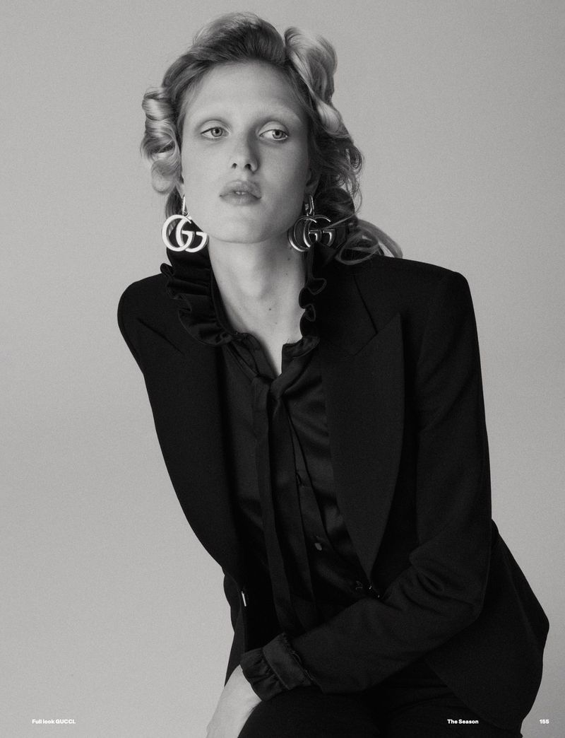 Elegant studio portrait of a woman with blonde hair, wearing a black blazer and statement earrings with a gray backdrop.