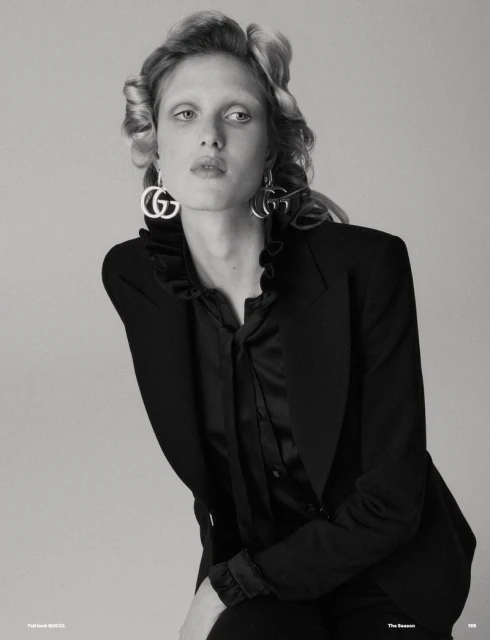 Elegant studio portrait of a woman with blonde hair, wearing a black blazer and statement earrings with a gray backdrop.