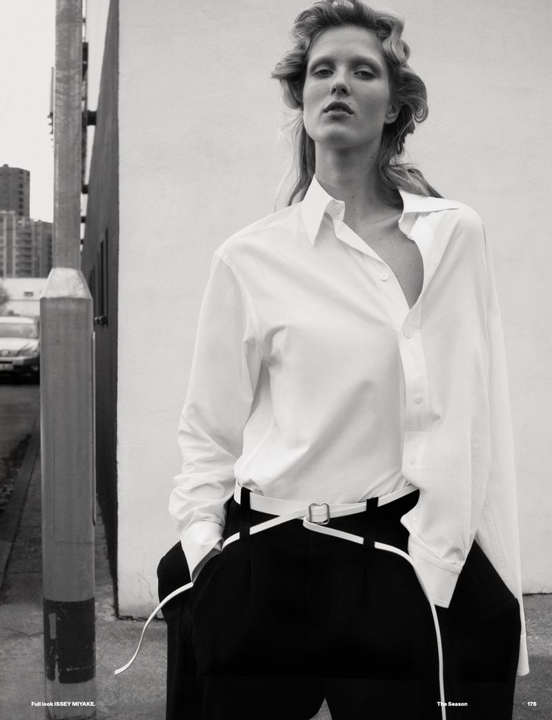 Editorial fashion shot of a blonde model in a white shirt and black pants with a white belt on a city street with buildings.