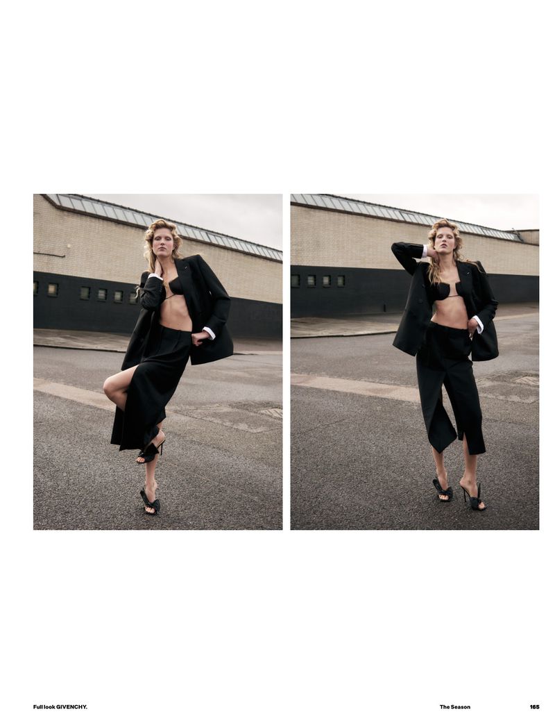 Fashion editorial shot in an urban environment features a model posing in a black blazer, bra top, and skirt with a tan background.