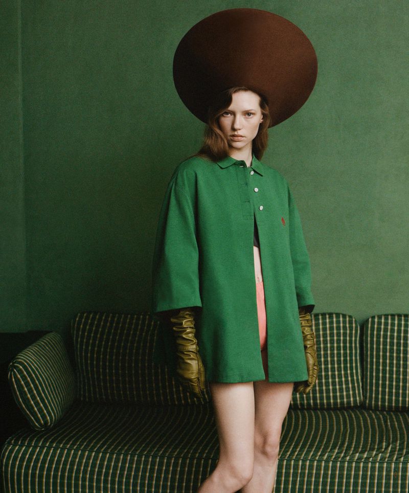 An adult female model wearing a green jacket and large brown hat poses in a studio in front of a couch and green wall.