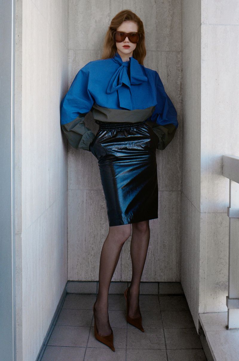 A fashionable woman stands in a corner wearing a color-blocked top, black shiny skirt, tights, brown pumps, and brown sunglasses.