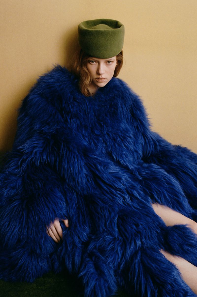 Portrait of a fashionable adult female in a large blue faux fur coat and olive hat sitting against a beige wall indoors in a studio.