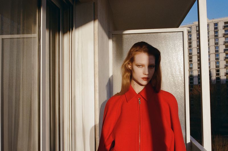 Model wearing a bright red coat on a sunny balcony with apartment buildings visible in the background