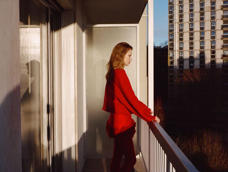 Adult woman in red outfit stands on a balcony overlooking a city building and dark green trees on a sunny day with clear sky.