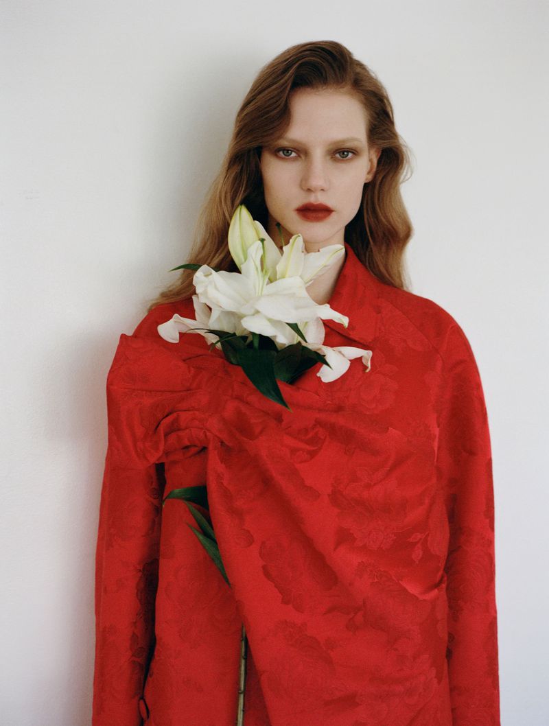 Indoor fashion portrait of a woman with long brown hair, red lipstick, and a red coat, holding a bouquet of white lilies.