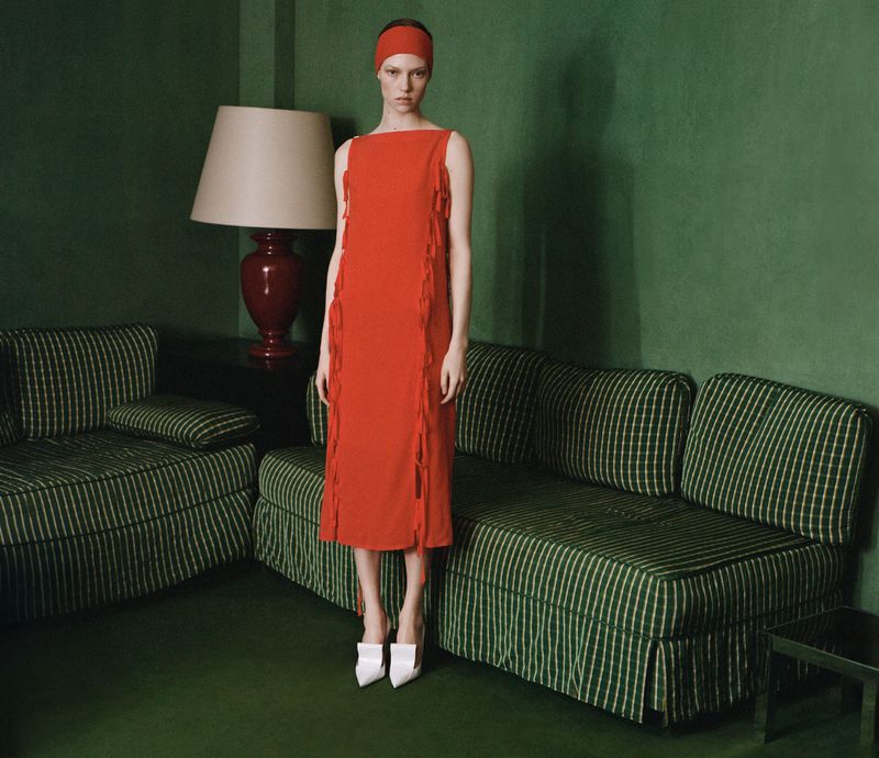 Model in red dress and headband standing in a green room with striped sofas and a large lamp