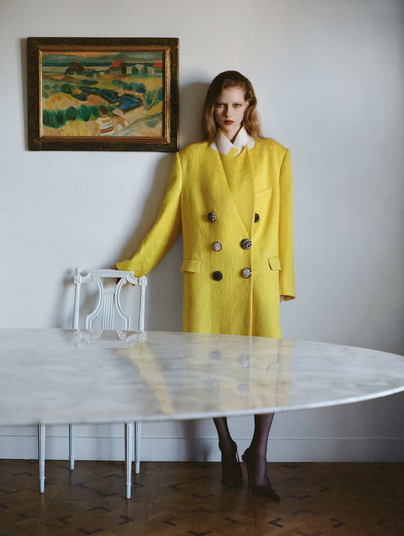 A fair-skinned model with blonde hair poses indoors next to a white marble table, wearing a yellow double-breasted blazer-dress.