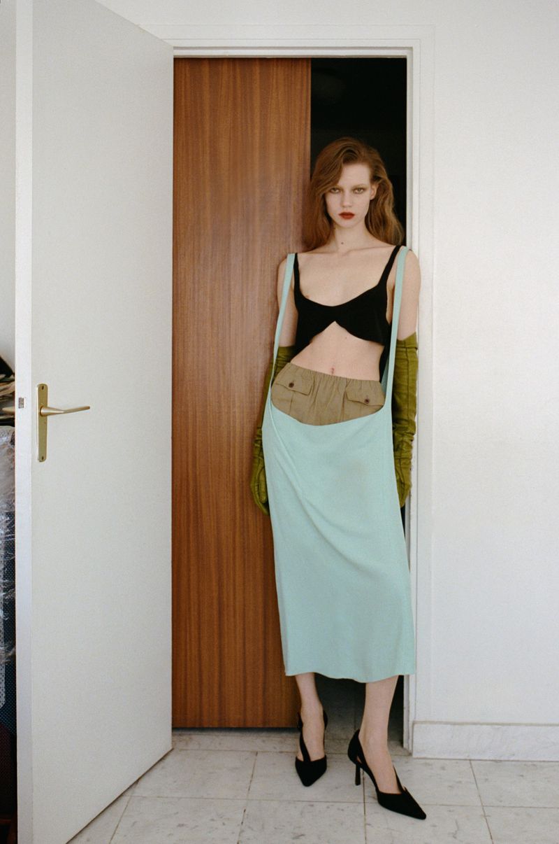 A red-haired fashion model poses in front of a partly open doorway in a black bra top and light-blue skirt with a tan utility belt.