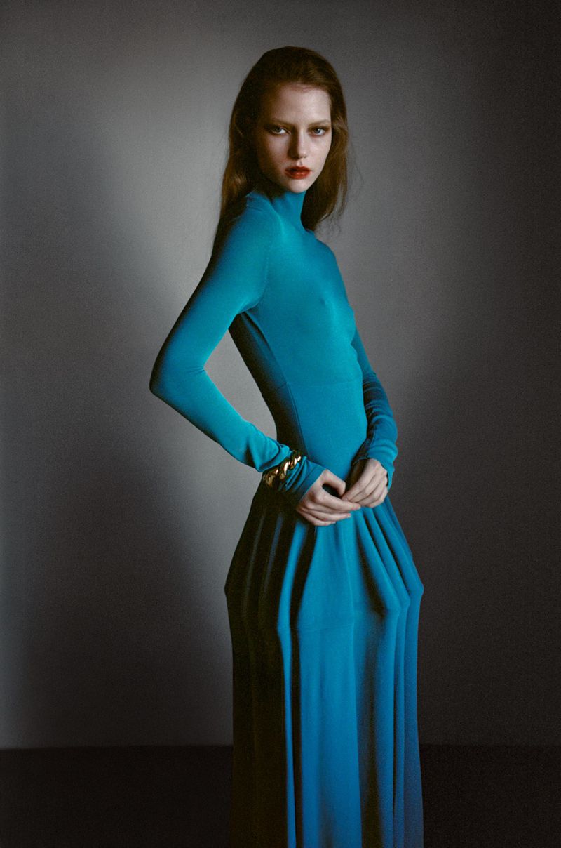 Fashion shot of a slim woman in a long-sleeved teal dress, standing in a photo studio with hands clasped and wearing gold bracelets.