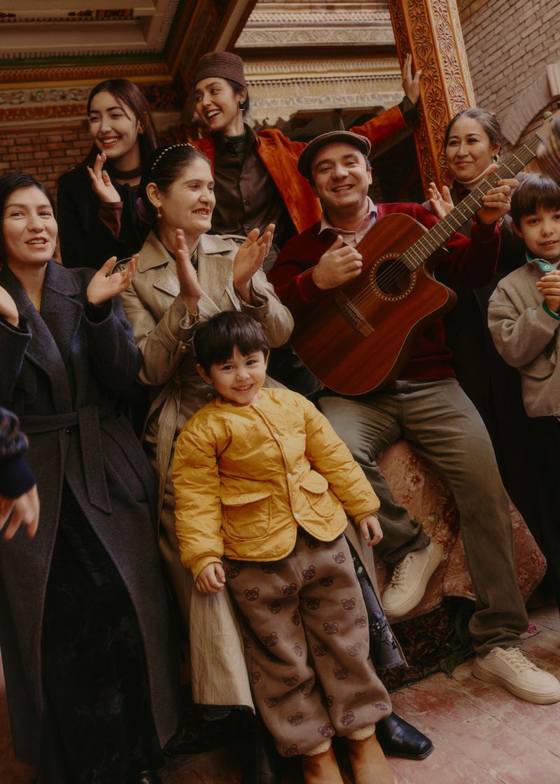 Group portrait of adults and children gathered indoors, man playing guitar, group smiling and clapping hands, traditional interior.