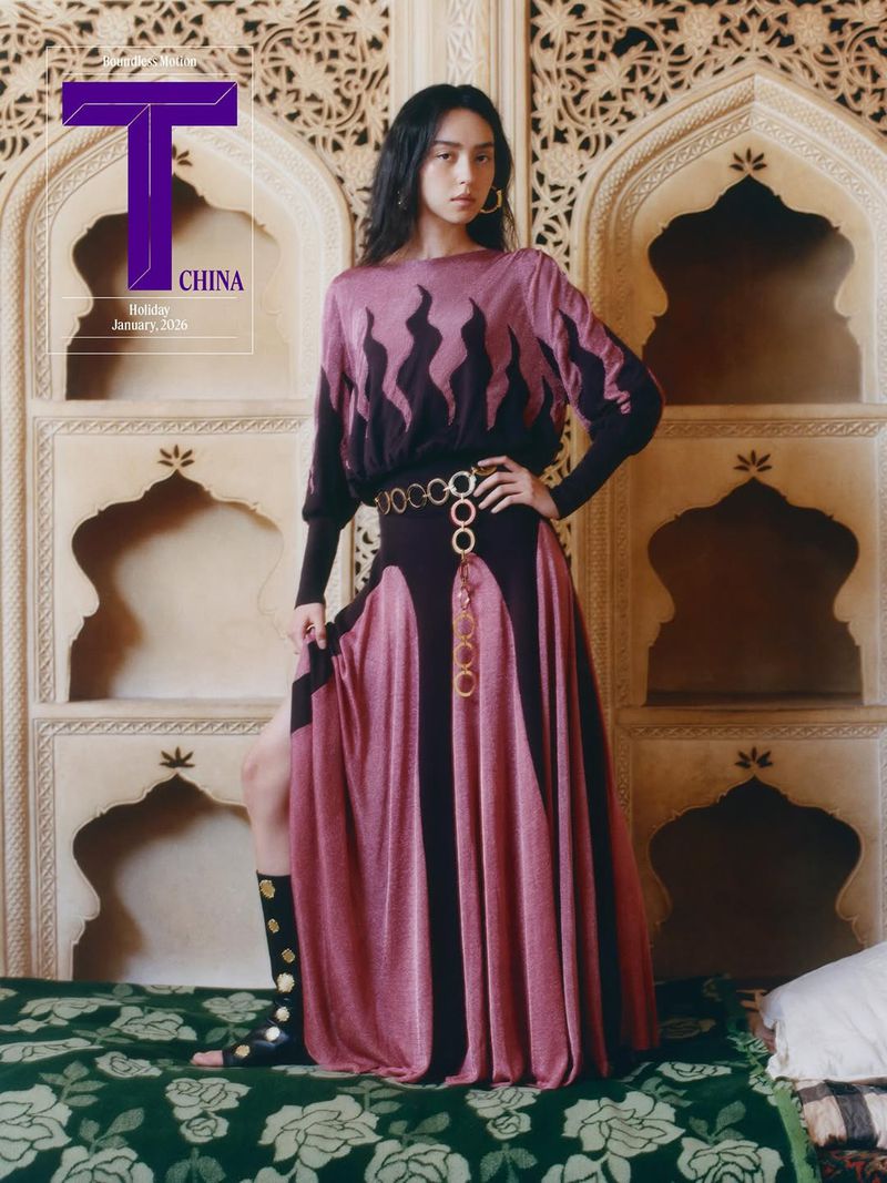 Fashion editorial photo featuring an adult female model in a long dress and black boots posing in front of an ornately decorated wall.