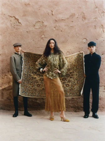 Editorial fashion shot of a woman in a patterned top and long skirt, holding a carpet with two male models in front of a textured wall.