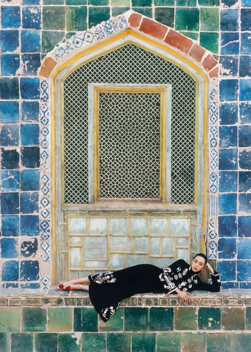 A woman in a black dress with white embroidery poses near a patterned window and tiled wall, bathed in soft, diffused natural light.