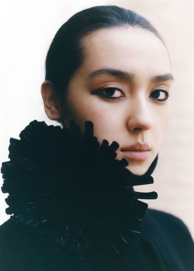 Close-up portrait of a young adult woman with prominent black eyeliner wearing a black textured collar, photographed in a photo studio.